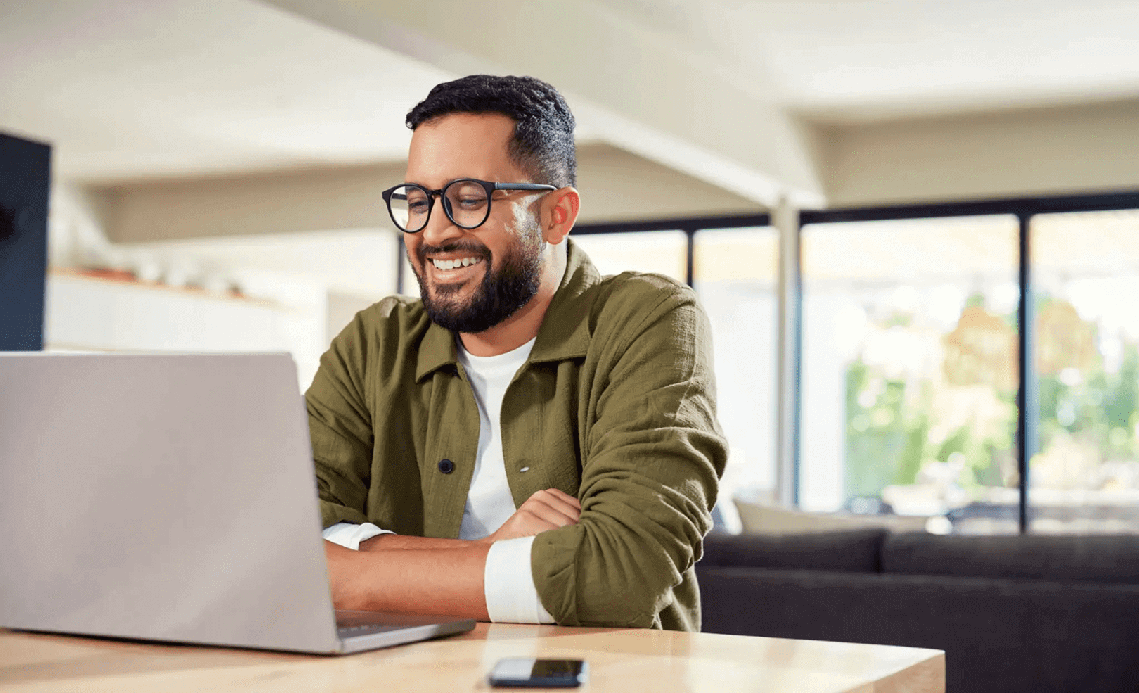 Man with glasses smiling while using a laptop at home.
